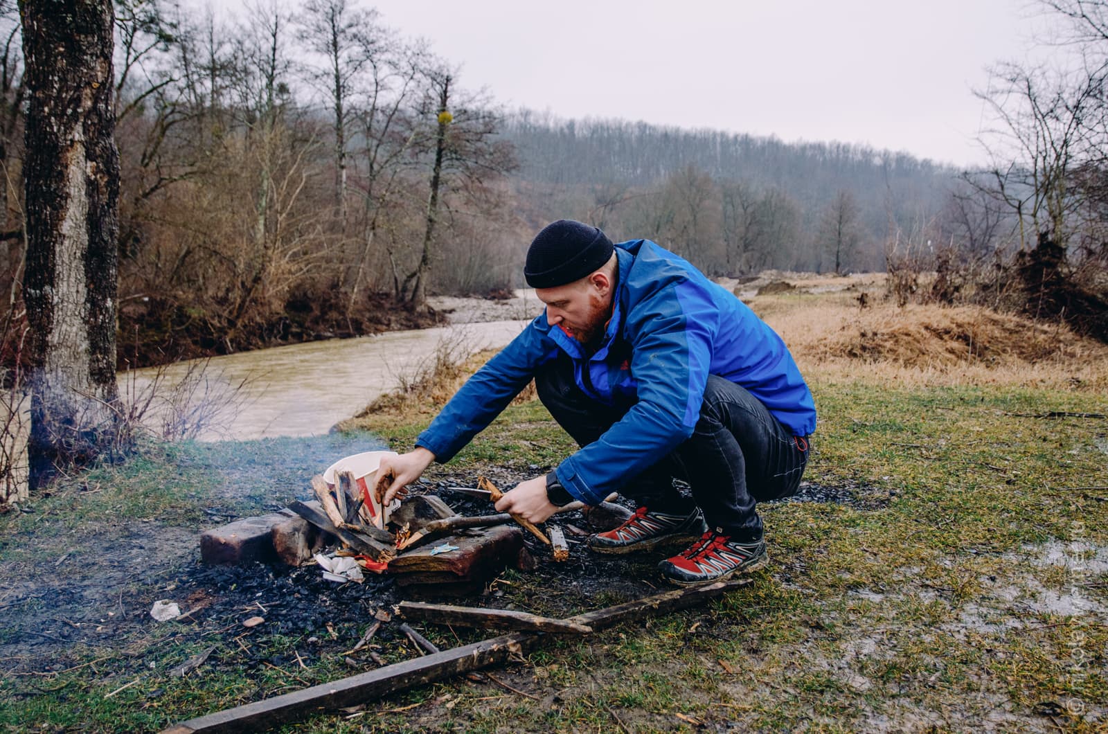 Человек в синей куртке сидит на корточках, разжигая костёр на берегу реки