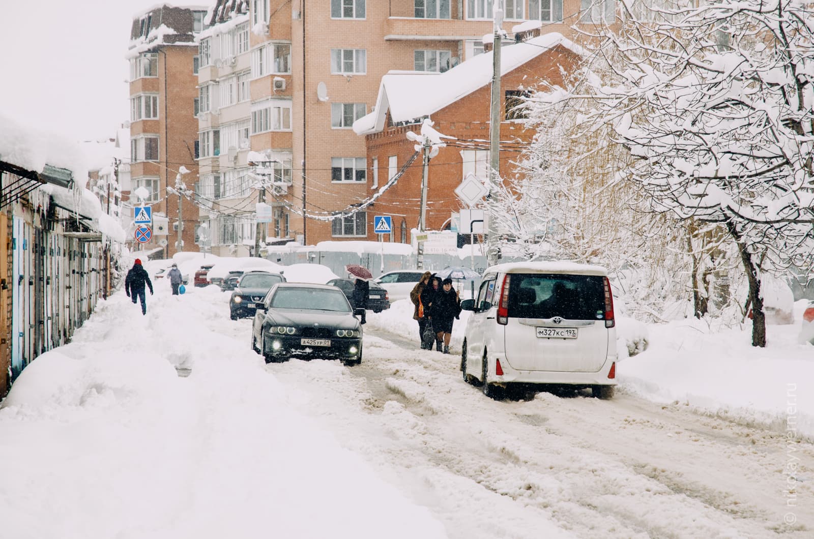 Узкая улица, занесённая снегом. Слева ряд гаражей, справа деревья, впереди кирпичные пятиэтажки. По снежным ухабам на проезжей части продираются автомобили