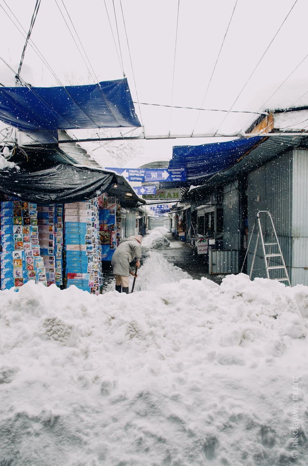 Большая стена снега, за ней длинный ряд ларьков и женщина чистит снег возле своего торгового места