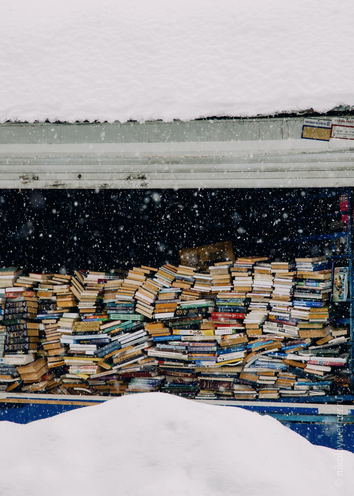 Стена снега на переднем плане, за ней прилавок, до верха забитый книгами, сверху снова снег, на крыше ларьк
