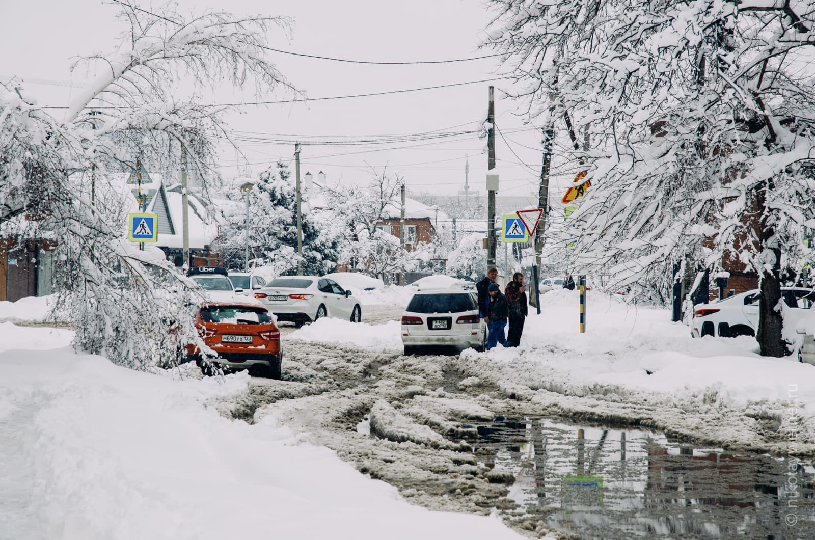 Узкая улица, на которой всё в снегу, но на проезжей части образовалсь большая лужа из подтаявшего снега