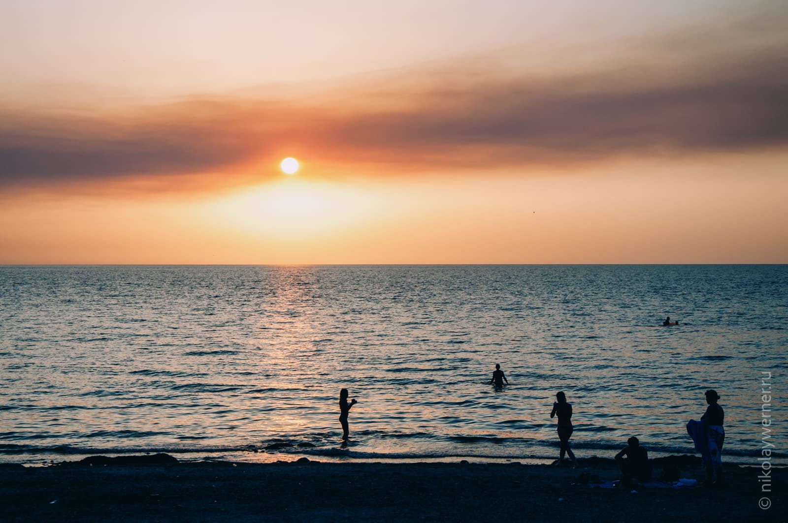 Закат. Вид с высокой набережной. Чёрная полоса берега, силуэты людей в воде и на берегу, спокойное море, шарик солнца на ладонь от горизонта и дымчатые облака.