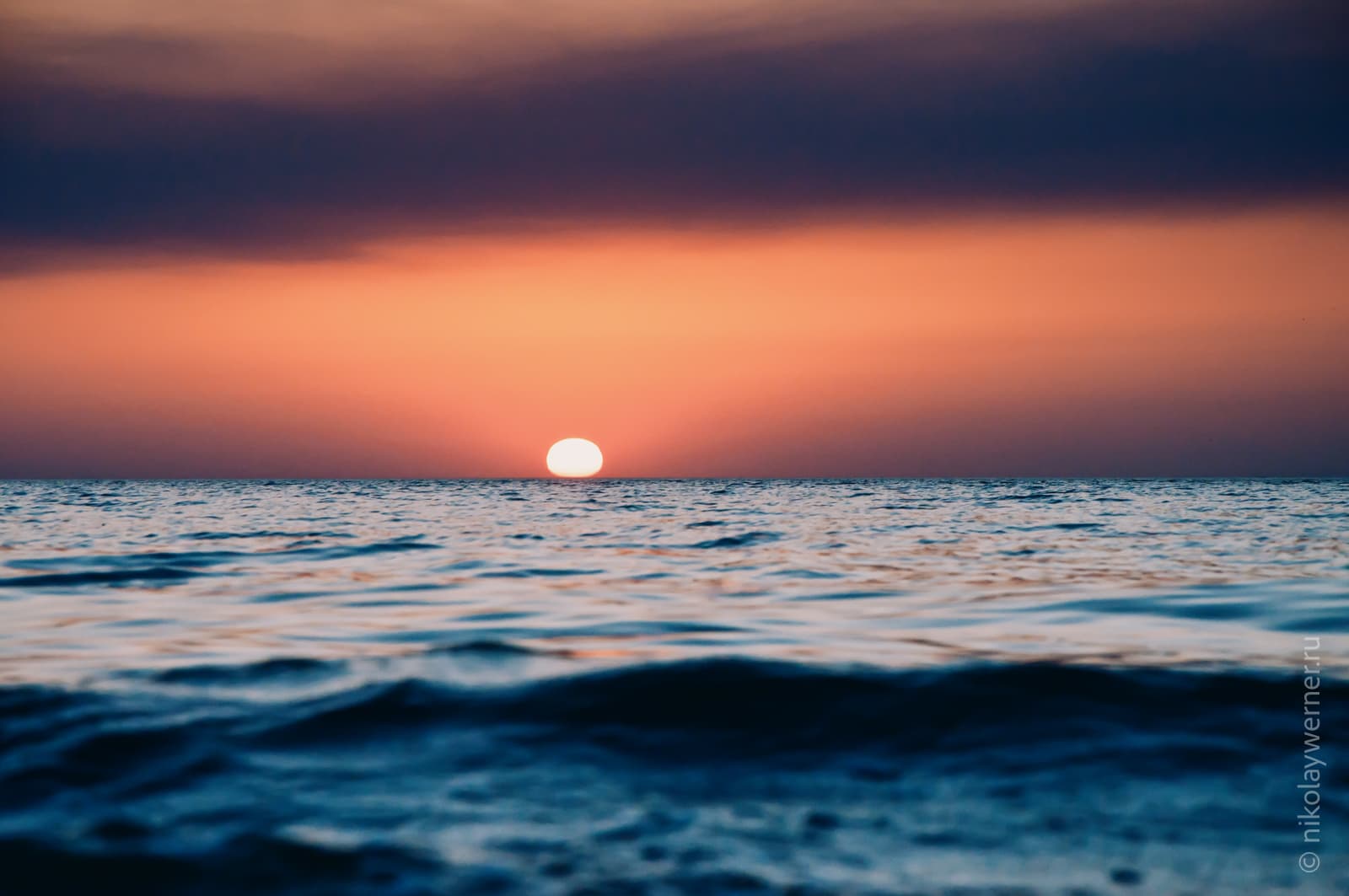 Закат. Солнце коснулось горизонта. Фотография от самой воды. На море лёгкая рябь, над горизонтом рыжее небо и тёмное дымчатое облако вверху.