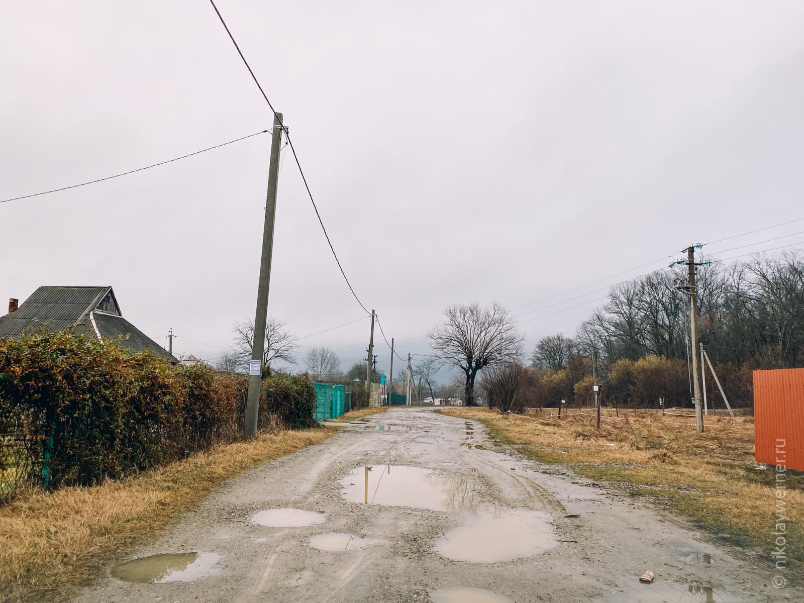 Грязная дорога с лужами. Пожухлая трава. Заросшая изгородь слева, поросший пустырь справа. Столбы, голые деревья дальше.