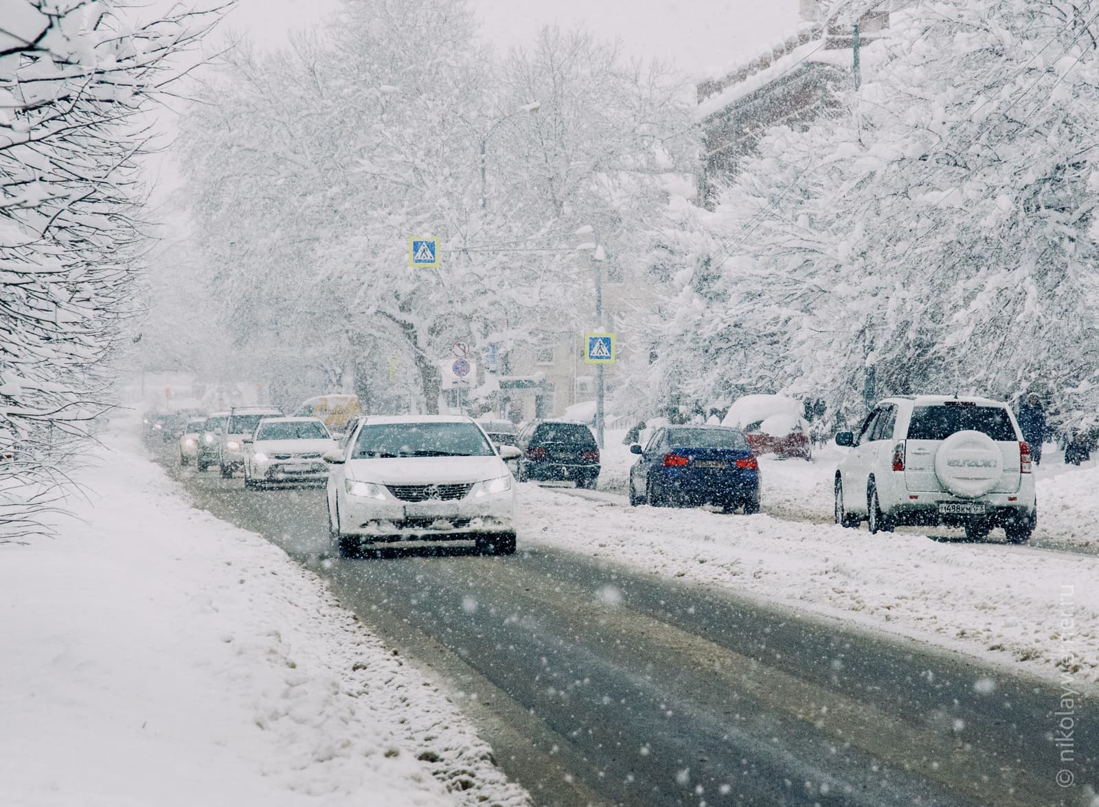 Чёрная полоса асфальта, вокруг всё в снегу: тротуары, деревья. Вереницей идут автомобили, сыплет снег.