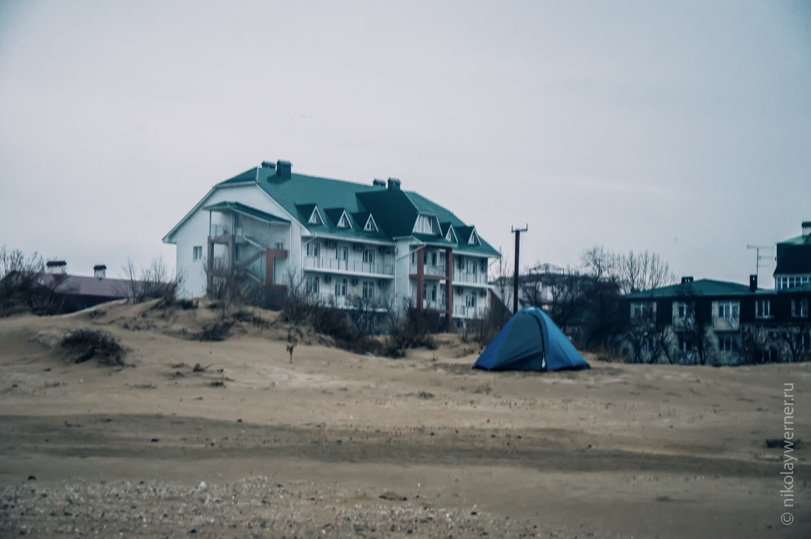 Синяя палатка среди песчанных дюн на пляже. Пасмурно. На заднем плане стоит трехэтажная гостиница.